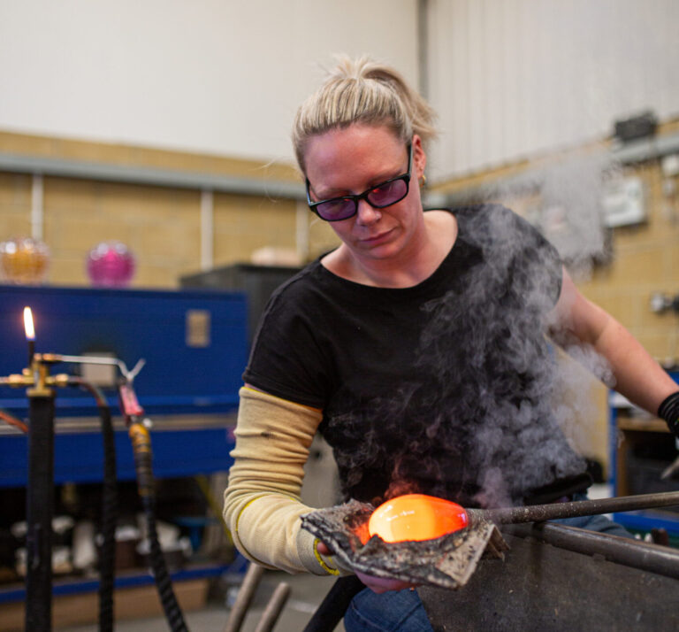 In this image you can see Katie sitting in the glass blowing bench, she is shaping a glass bubble with some newspaper. Katie has blonde hair, which is tied back and is wearing purple UV safety glasses and a kevlar sleeve.