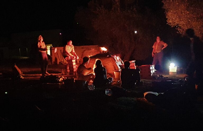 A group of people in the dark of night, labouring under the glow from a man-made furnace, the orange hue illuminates their faces as they stoke the fire and sit around the furnace watching the stoking.